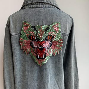 May include: A gray denim jacket with a large, colorful sequined cat face design on the back. The cat has green, pink, and black sequins and a red mouth.