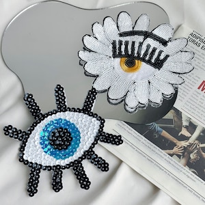 以下が含まれることがあります： アイロン接着できる刺繍パッチ2枚。1枚は白いデイジーに黒い目とまつげ、もう1枚は青い目に黒いまつげと白と黒のキラキラしたものがついています。