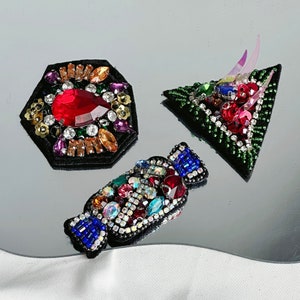Peut inclure: Trois patchs brodés colorés avec des strass et des perles. Les patchs sont en forme de carré, de bonbon et de triangle avec des pointes.