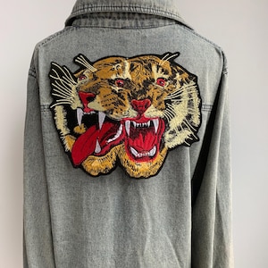 May include: A gray denim jacket featuring a large embroidered tiger patch on the back. The tiger patch has a detailed design with shades of brown, gold, red, and black, showcasing an open mouth with visible teeth and tongue.
