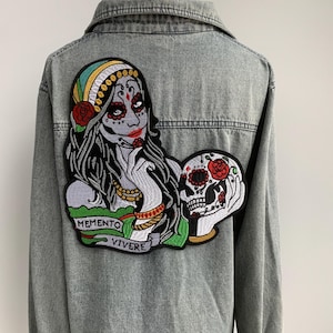 Könnte beinhalten: Eine Jeansjacke mit einem gestickten Aufnäher, der eine Frau mit einem Schädel und dem Text "Memento Vivere" zeigt.
