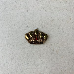 Op de afbeelding: Een goudkleurige kroon met een rode edelsteen in het midden. De kroon is versierd met gouden kralen en druppelvormige edelstenen. Deze decoratieve kroon is geschikt voor het verfraaien van kleding of accessoires.