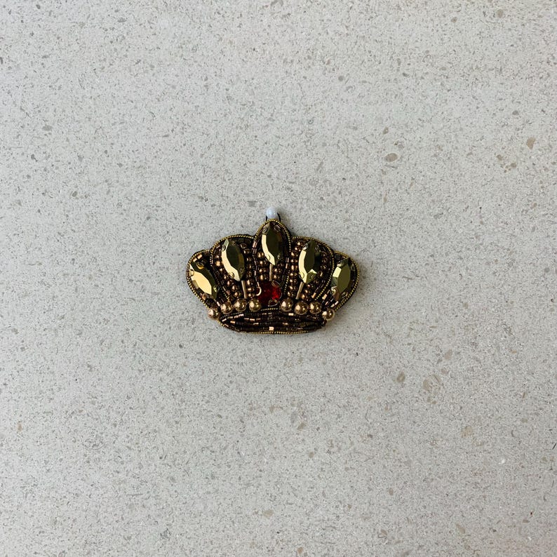 Op de afbeelding: Een goudkleurige kroon met een rode edelsteen in het midden. De kroon is versierd met gouden kralen en druppelvormige edelstenen. Deze decoratieve kroon is geschikt voor het verfraaien van kleding of accessoires.