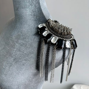 May include: A black and silver shoulder piece with a fringe of chains. The piece is decorated with black and clear rhinestones.
