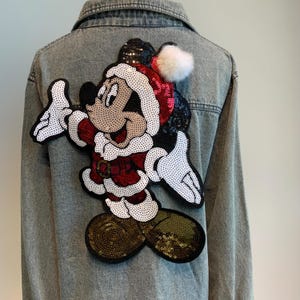 Könnte beinhalten: Eine Jeansjacke mit einem Pailletten-Mickey-Mouse-Aufnäher auf der Rückseite. Mickey trägt einen roten und weißen Weihnachtsmann-Anzug und eine weiße Pelzmütze.