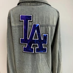Op de afbeelding: Grijze spijkerjas met een groot blauw en wit pailletten "LA" logo op de rug. De jas heeft lange mouwen en een klassiek knoopontwerp. Het logo is een opvallend detail.