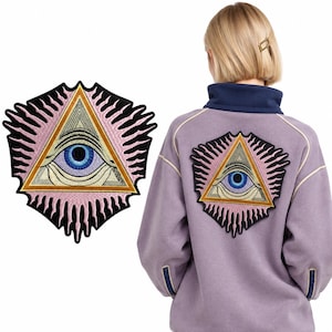 May include: An embroidered patch featuring a central blue eye inside a golden triangle, surrounded by pink radiating lines and a black border. The same patch is shown applied to the back of a light purple fleece jacket with a dark blue collar.