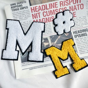 May include: Three felt patches in the shape of the letter "M". One is white with black outlines, one is yellow with black outlines, and one is white with black outlines and a hashtag symbol. The patches are on a printed page with text.