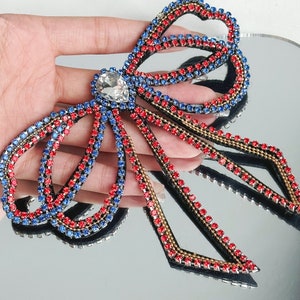 Könnte beinhalten: Eine große Schleife aus roten, blauen und goldenen Strasssteinen mit einem großen, klaren Strassstein in der Mitte. Die Schleife besteht aus Stoff und Strasssteinen und ist zum Annähen oder Aufbügeln auf Kleidung oder andere Gegenstände gedacht.