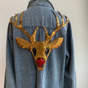 Puede incluir: Chaqueta vaquera azul claro con un gran diseño de reno con lentejuelas doradas en la espalda. El reno tiene ojos negros y una nariz roja con lentejuelas. La chaqueta tiene cierre de botones y cuello clásico.