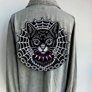 May include: A grey denim jacket featuring a large embroidered patch on the back. The patch depicts a black cat face centred within a white spider web, wearing a purple spiked collar, and framed by a white chain link border design.