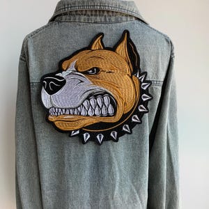Puede incluir: Chaqueta vaquera azul claro con un gran parche bordado de la cabeza de un perro enfadado en la espalda. El parche presenta colores marrón, gris y negro con un collar de púas.
