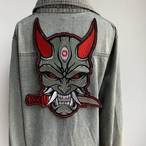 Peut inclure: Veste en jean bleu clair avec un grand écusson brodé d'un masque Oni japonais. Le patch présente un masque gris avec des cornes et des yeux rouges, une bouche souriante aux dents blanches et un couteau.