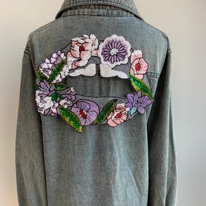 May include: A gray denim jacket with a large embroidered floral design on the back. The design features a variety of flowers in shades of pink, purple, and green.