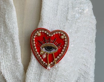 Op de afbeelding: Rode vilt hartvormige broche met een parel oog en gouden accenten. Het oog heeft een zwarte pupil en witte highlights. De broche heeft drie kristal traan versieringen.