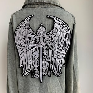 Puede incluir: Chaqueta vaquera gris con un parche bordado grande en la espalda. El parche presenta un diseño detallado de un ángel guerrero con una espada y grandes alas, en blanco y negro.