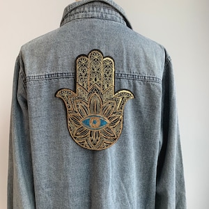 以下が含まれることがあります： 青い目を中心に、金と黒の刺繍が施されたハンズのデニムシャツです。