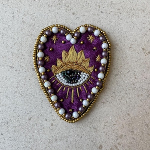 Op de afbeelding: Een hartvormige, geborduurde patch met een paarse fluwelen achtergrond. De patch heeft een goudkleurig oogontwerp met een zonnestraal, omgeven door gouden kralen en witte en paarse parels. De patch is ongeveer 7,6 cm breed.