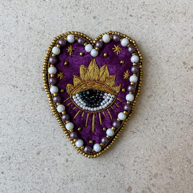 Op de afbeelding: Een hartvormige, geborduurde patch met een paarse fluwelen achtergrond. De patch heeft een goudkleurig oogontwerp met een zonnestraal, omgeven door gouden kralen en witte en paarse parels. De patch is ongeveer 7,6 cm breed.