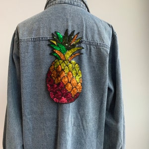 Può includere: Una camicia di jeans blu con una toppa di ananas di paillettes colorate sul retro.