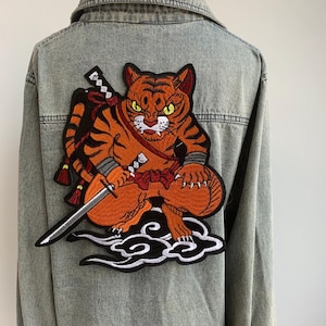 May include: A light-wash denim jacket featuring a large embroidered patch of an orange tiger in a warrior pose, holding a sword. The tiger is set against a white cloud design. The jacket has a classic collar and button-up front.