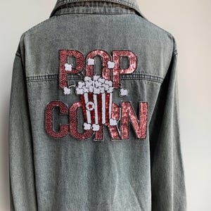 Könnte beinhalten: Eine Jeansjacke mit einem pinkfarbenen und weißen Pailletten-Aufnäher, auf dem "POPCORN" steht und in der Mitte ein Eimer Popcorn abgebildet ist.