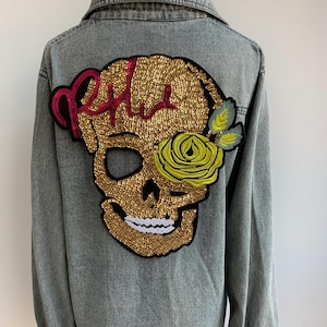 May include: A gray denim jacket with a large gold sequined skull and a yellow rose embroidered on the back. The skull has a red and pink sequined design on the top.