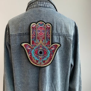 May include: A blue denim jacket with a large embroidered patch of a hamsa hand with an eye in the center. The hamsa hand is a symbol of protection and good luck.