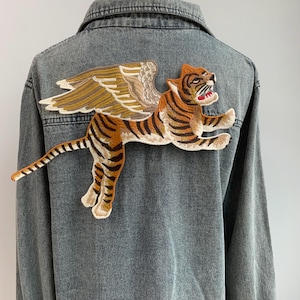 Może przedstawiać: Dżinsowa kurtka z dużą haftowaną naszywką przedstawiającą tygrysa z skrzydłami. Tygrys jest pomarańczowy i czarny ze skrzydłami brązowymi.