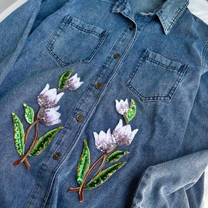 Może przedstawiać: Dżinsowa kurtka z trzema haftowanymi naszywkami białych tulipanów z zielonymi liśćmi. Tulipany są wykonane z cekinów.