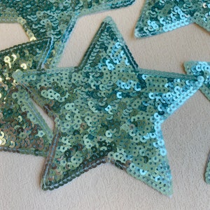 May include: Five teal blue star-shaped fabric patches covered in sequins. The stars are sewn with white thread.