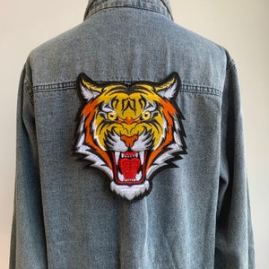 Op de afbeelding: Een spijkerjack met een grote geborduurde tijgerpatch op de rug. De tijger is oranje, geel en zwart met een witte bek en tanden.
