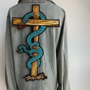 May include: Gray denim jacket featuring a large embroidered patch on the back. The patch depicts a teal snake coiled around a wooden cross with a red tongue. The cross is brown with a gold border.