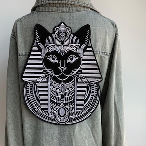 Könnte beinhalten: Ein schwarz-weißer, gestickter Aufnäher einer Katze im Stil der altägyptischen Pharaonen, befestigt an einer hellblauen Jeansjacke. Die Katze hat eine detaillierte Kopfbedeckung und einen Kragen mit aufwendigen weißen Nähten auf schwarzem Grund.
