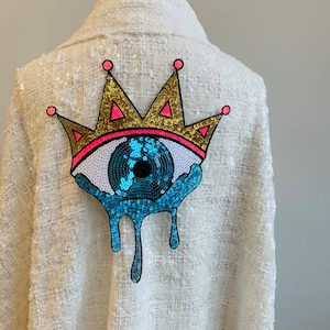 May include: A white sequined jacket with a large blue eye and gold crown applique. The eye is dripping with blue sequins.