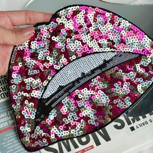 May include: A pink and silver sequined patch in the shape of a pair of lips. The lips are outlined in black and have a white, shiny, textured surface.