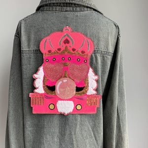 以下が含まれることがあります： 背中に大きなピンクのスパンコールパッチが付いたグレーのデニムジャケット。パッチには、王冠、ハート型のサングラス、ガムバブルを持つ漫画のキャラクターが描かれています。キャラクターは白い髪と金色のアクセントを持っています。