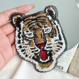 May include: A close-up of a tiger face embroidered with sequins and beads. The tiger's face is a mix of black, white, gold, and red.