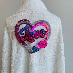 Pode incluir: Casaco branco de lantejoulas com um grande remendo em forma de coração rosa, roxo e prateado que diz "Love" em letras rosa. O remendo em forma de coração também tem um lábio rosa e um diamante prateado.