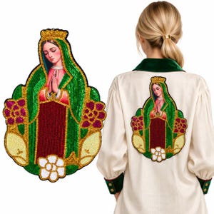 Puede incluir: Un parche grande de lentejuelas con la imagen de la Virgen de Guadalupe con manto verde, vestido rojo y corona dorada, rodeada de flores rosas. El parche se muestra aplicado en la parte posterior de una camisa color crema de manga larga.