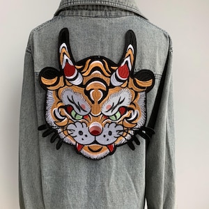 Può includere: Giacca di jeans grigia con una grande toppa ricamata a forma di testa di tigre sul retro. La toppa presenta un design dettagliato con colori arancione, nero, bianco, rosso e verde. La giacca ha un colletto classico e una chiusura con bottoni.