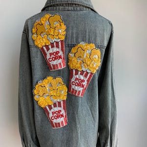 Könnte beinhalten: Eine graue Jeansjacke mit drei Popcorn-Patches. Die Patches sind aus Pailletten und haben rote und weiße Streifen mit dem Schriftzug "POP CORN" in Weiß.