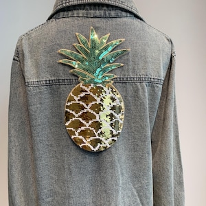 Può includere: Una giacca di jeans con una grande toppa di ananas di paillettes dorate e verdi sul retro.
