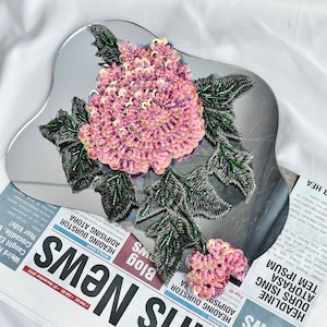 May include: A pink and green floral applique with sequins. The applique is on a mirrored surface.