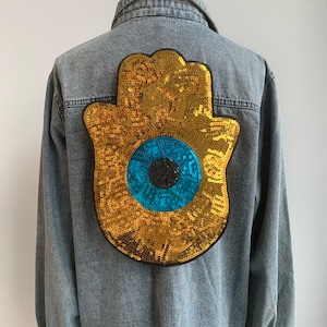 以下が含まれることがあります： 青い邪悪な目を中心に、金色のスパンコールでできたハmsaの手がついたデニムジャケットです。