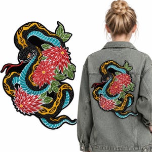 Puede incluir: Un parche bordado que muestra una serpiente negra con escamas amarillas y turquesas, enroscada alrededor de dos grandes flores de crisantemo rojas con hojas verdes, sobre la espalda de una chaqueta vaquera gris.