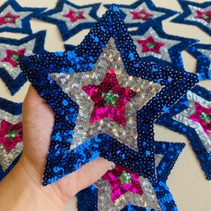 Puede incluir: Primer plano de una gran estrella con lentejuelas en tonos azules, plateados y rosas. La estrella tiene un diseño en capas con una estrella rosa central y una capa intermedia plateada, todo delineado con lentejuelas azules. Varias estrellas similares son visibles en el fondo.