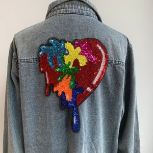 Könnte beinhalten: Hellblaue Jeansjacke mit einem großen paillettenbesetzten Herzdesign auf dem Rücken. Das Herz ist rot mit farbenfrohen tropfenden Akzenten in Blau, Orange, Grün, Gelb und Rosa. Die Jacke hat einen klassischen Kragen und eine Knopfleiste.