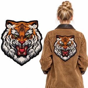 May include: An embroidered tiger head patch featuring orange, white, and black fur details with green eyes and an open mouth showing teeth. The patch is shown separately and applied to the back of a brown jacket with a dark collar.