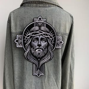 Può includere: Giacca di jeans grigia con una grande toppa ricamata sul retro. La toppa presenta un'immagine dettagliata in bianco e nero di Gesù Cristo con una corona di spine, su un disegno a forma di croce.